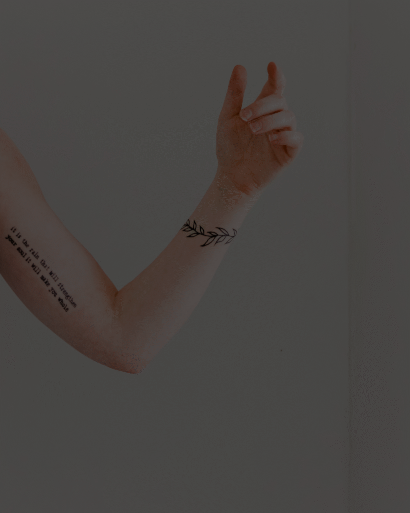Bras levé d’une personne avec deux tatouages : une branche feuillue autour du poignet et une citation en lettres noires sur l’avant-bras.