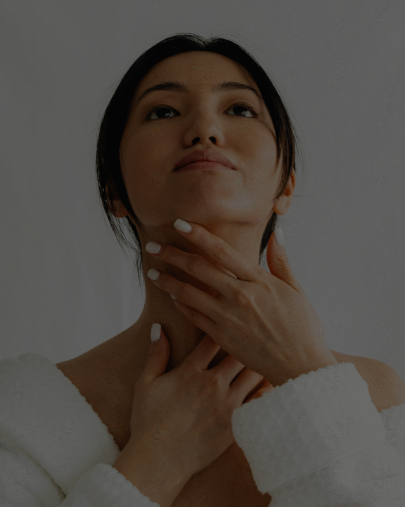 Femme en peignoir blanc touchant délicatement son cou et son menton, posant avec une expression sereine devant un fond clair