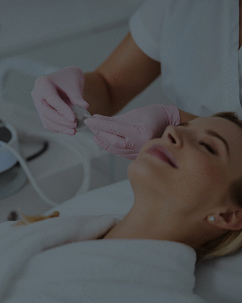 Une femme détendue allongée dans une clinique esthétique reçoit un soin du visage. Une estheticienne en gants roses prépare l’appareil de traitement connecté à une machine esthétique.
